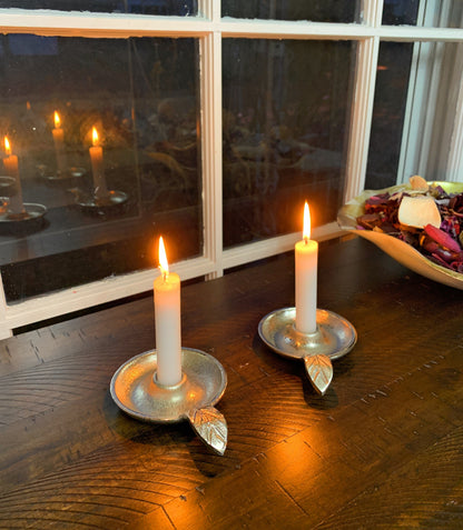 Two silver leaf-handle candle holders with lit tapers on a wooden table; holders sold individually